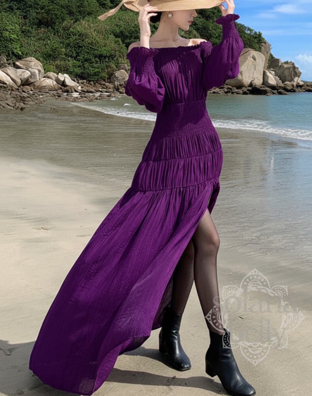 海辺でガラベーヤワンピースを着たモデルが帽子を持ち上げてポーズ。紫の生地が光を受けて揺れ、ベリーダンスのレッスン着や衣装に重ねても使いやすいシルエットが分かる写真。