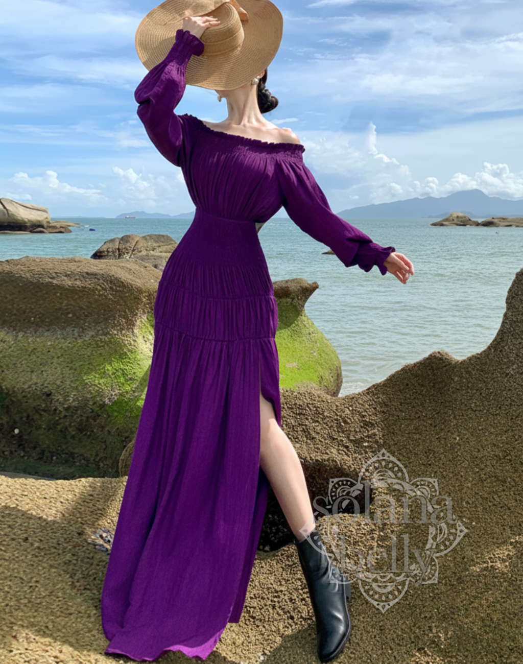 紫のガラベーヤワンピースを着たモデルが岩場で片足を軽く上げて後ろ向きに立つ姿。裾が風で流れ、ベリーダンスのレッスン着や衣装の上に羽織っても美しく見える動きが伝わる構図。