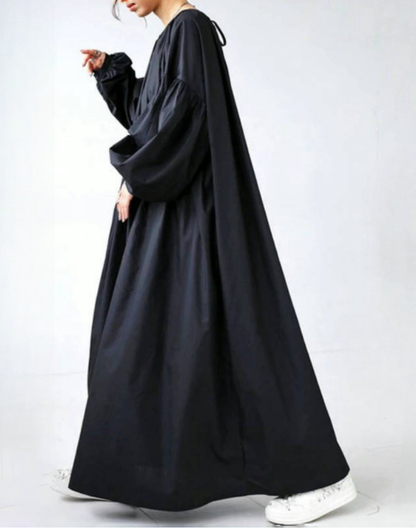 黒のガラベーヤワンピースを着たモデルが横向きで歩く姿を撮影。ハリのある生地が足元まできれいに広がり、ベリーダンスの衣装やレッスン着の上からも軽く羽織れるゆったりとしたシルエットが分かる構図。